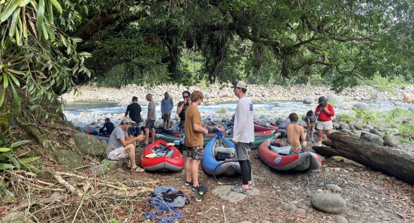 A group of people and watercraft gather on the shore of a river under a large tree